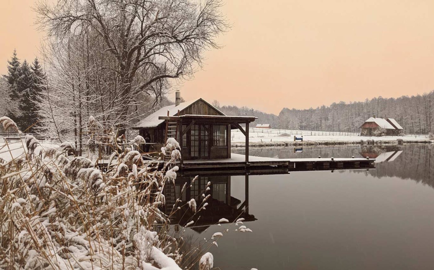 Zagroda Ojrzanów zimą, staw i drewniany domek na stawie. W tle budynek z sauną